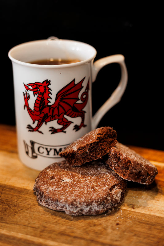 Mug with a red Welsh dragon design on a wooden surface with Dragons Breath Welsh Cakes in front of it.