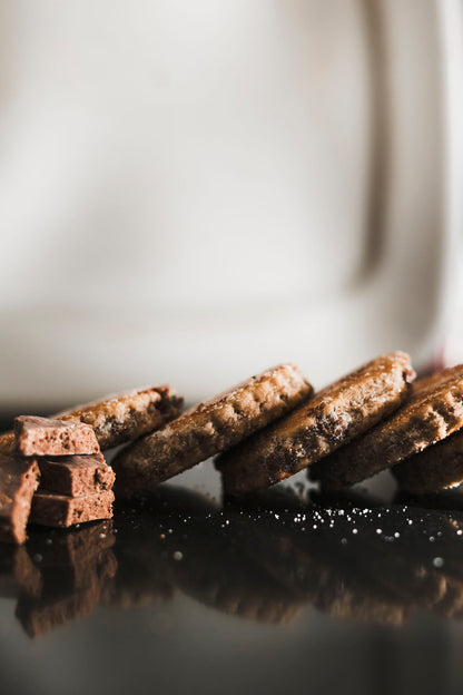 Close up of Dragonstone Bakes chocolate chip welsh cakes lying down next to a chunk of dairy milk chocolate on a dark reflective surface.