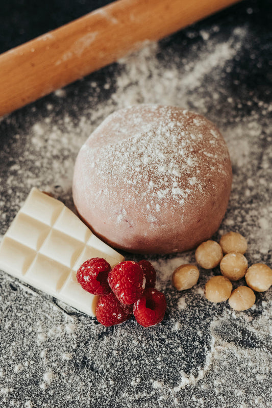 A ball of White Wizard Welsh cake dough with raspberries, a white chocolate bar, and macadamia nuts on a floured surface.